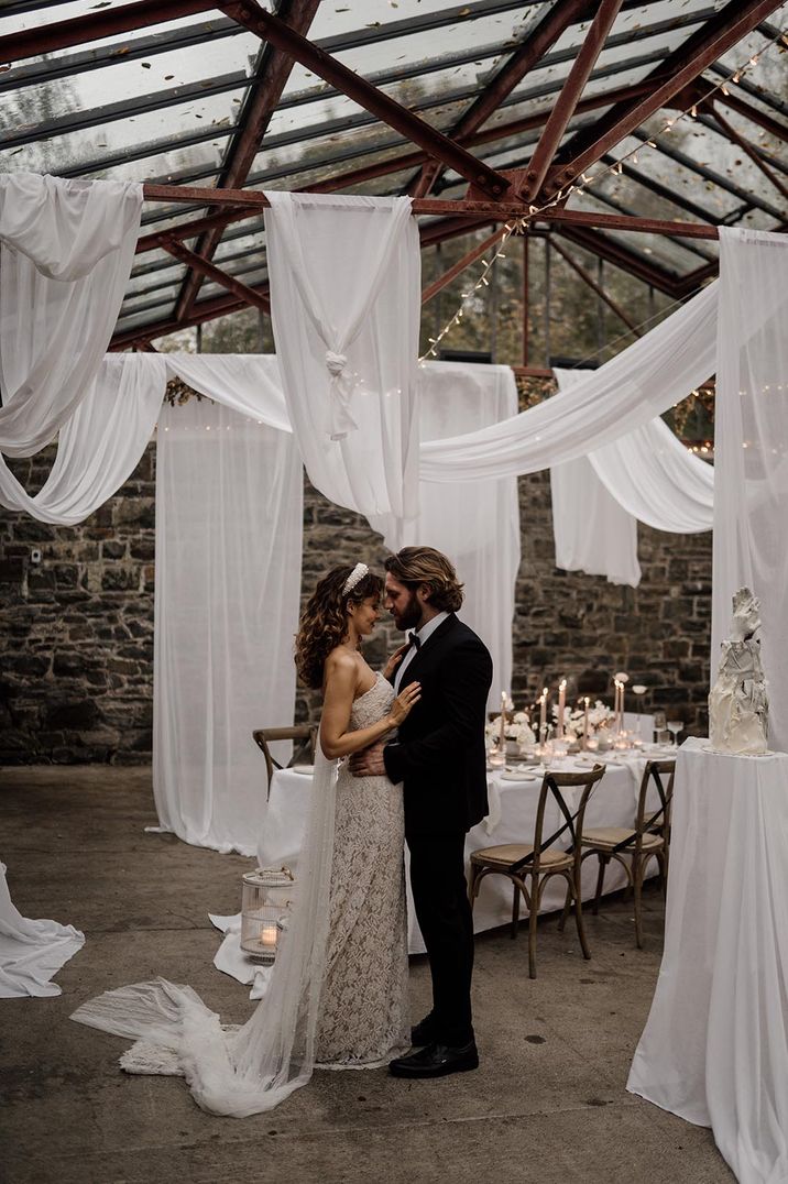 Bride and groom standing in the reception room of the glasshouse part of Plas Dinam Country House with white wedding ceiling drapes and fairy lights 