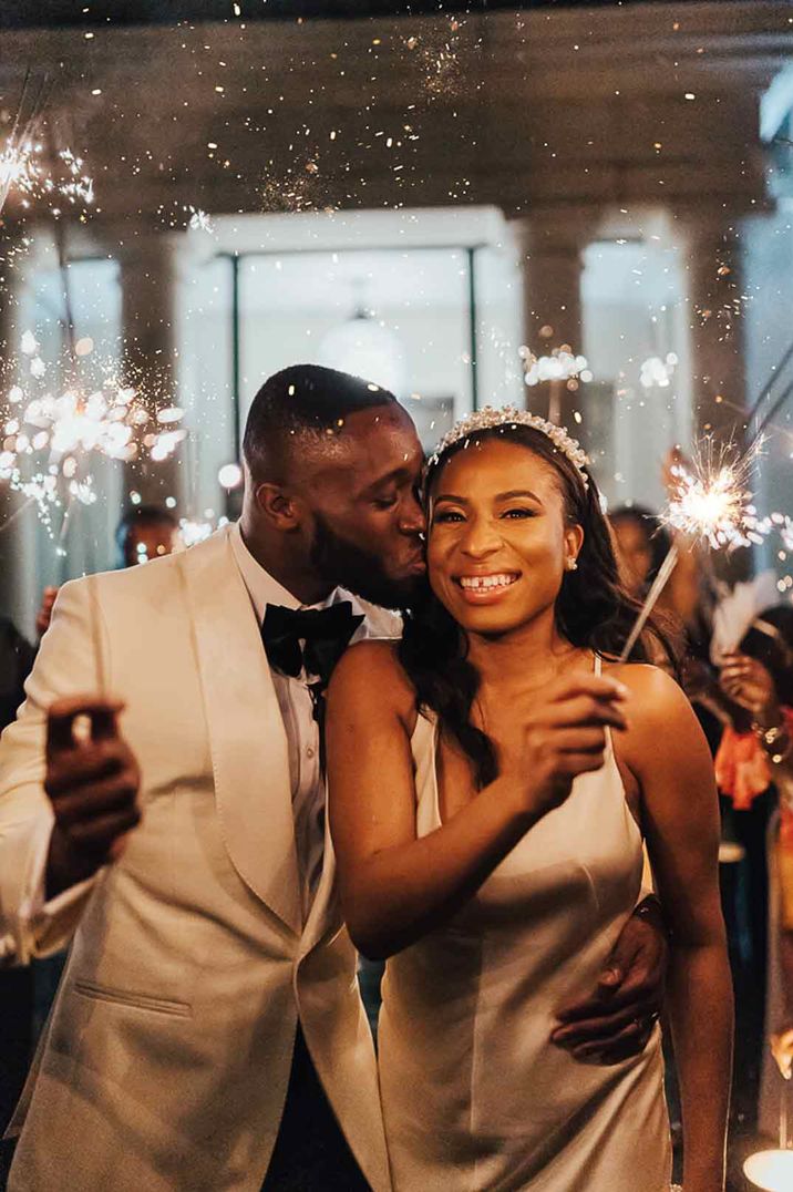 Bride in sleeveless satin wedding dress and pearl headband standing with groom in white tuxedo and black bow tie doing wedding sparkler exit 