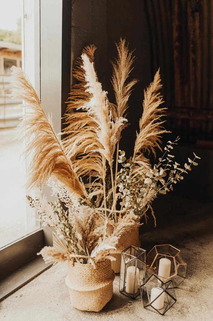 Pampas grass and eucalyptus wedding flower arrangements in wicker vases next to pillar candles in geometric glass candle holders 