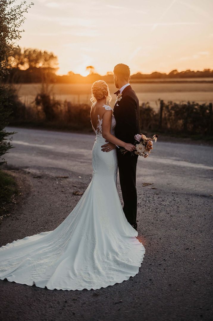 The bride wears a Pronovias wedding dress with her hair in an updo with the groom looking out at the sunset 