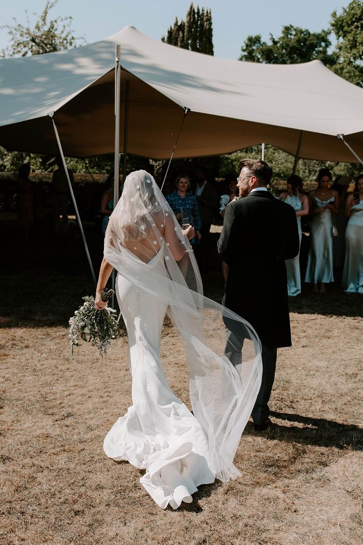 Bride in floral applique wedding veil walking with the groom to their marquee reception 