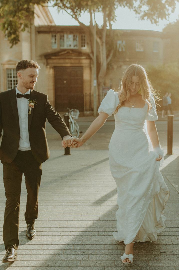 Bride in puff sleeve wedding dress walks during golden hour holding hands with the groom in black tie 