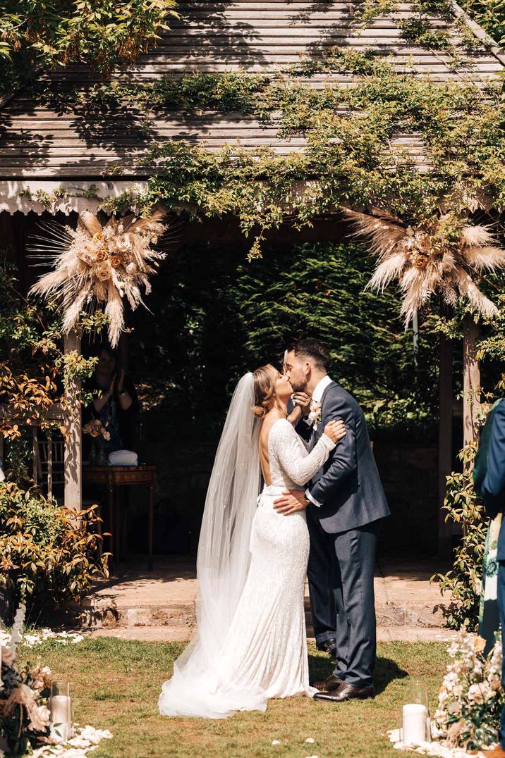 Bride in open back sequin wedding dress and groom in dark suit kissing at Pennard House with pampass press floral arrangements