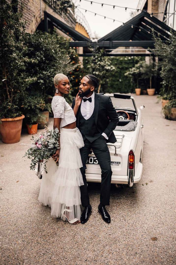 Bride in bridal two piece set with crop top and full tulle layered skirt standing with groom in classic black tuxedo 