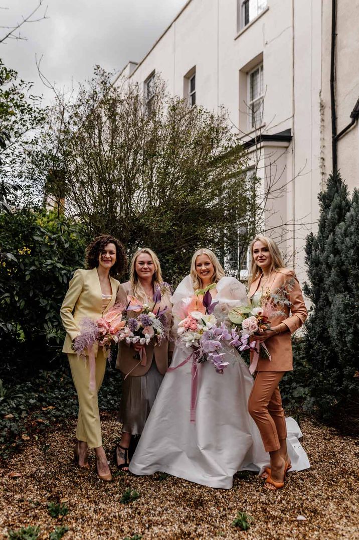 Bride in dramatic satin wedding dress with large puff sleeves standing with bridesmaids in mismatched pastel satin bridesmaid suits - hen party ideas