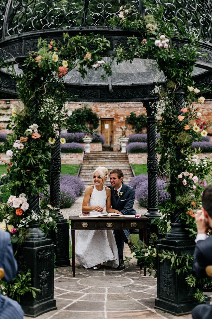 Bride in halterneck wedding dress and colourful bouquet and groom signing the register