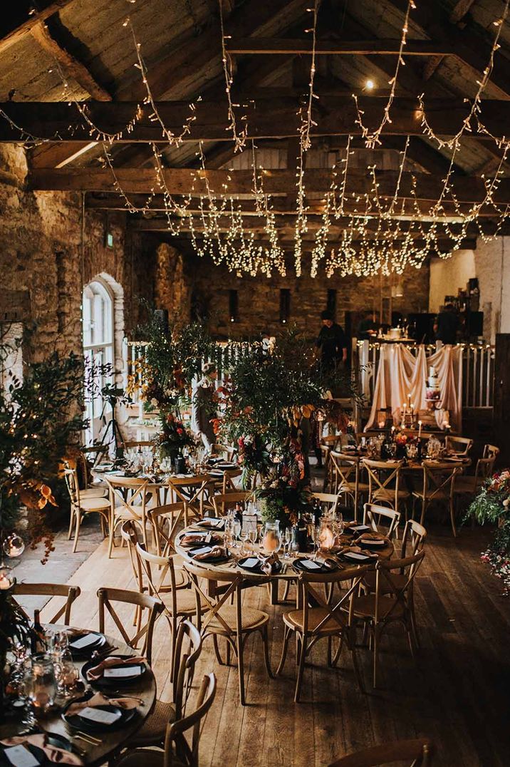 Autumnal themed wedding reception room in Askham Hall with wooden tables, wooden chairs, suspended fairy lights decor and wedding drapes 