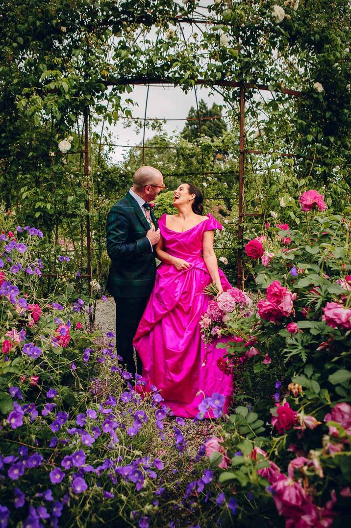 Bride in bright pink Vivienne Westwood wedding dress and groom in dark plaid grooms suit standing amidst colourful flowers at Middleton Lodge Estate wedding venue 