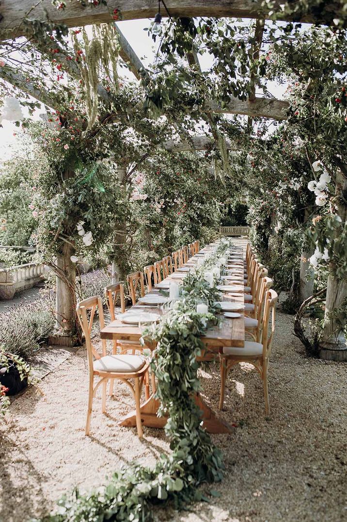 Al fresco wedding breakfast with wooden banquet table covered in waterfall foliage table runner with white pillar candles 