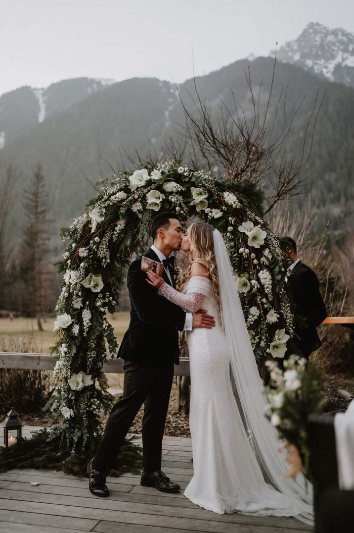 bride and groom kissing during the outdoor wedding ceremony with white and green floral arch 