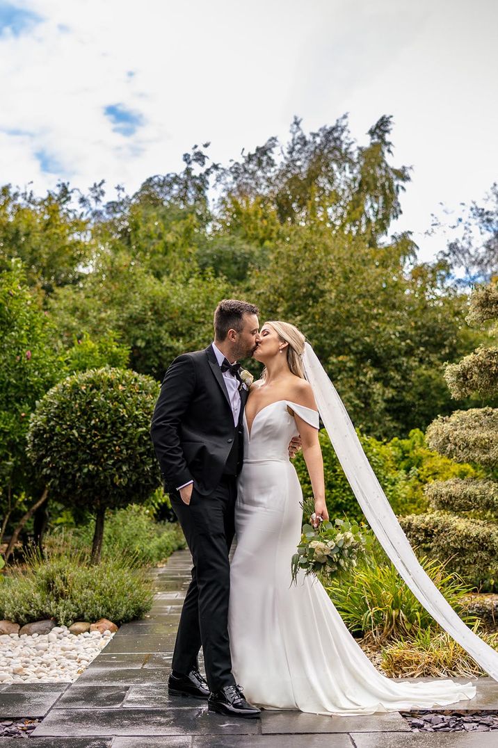 Bride in classic white wedding dress and veil kissing groom in black tie 