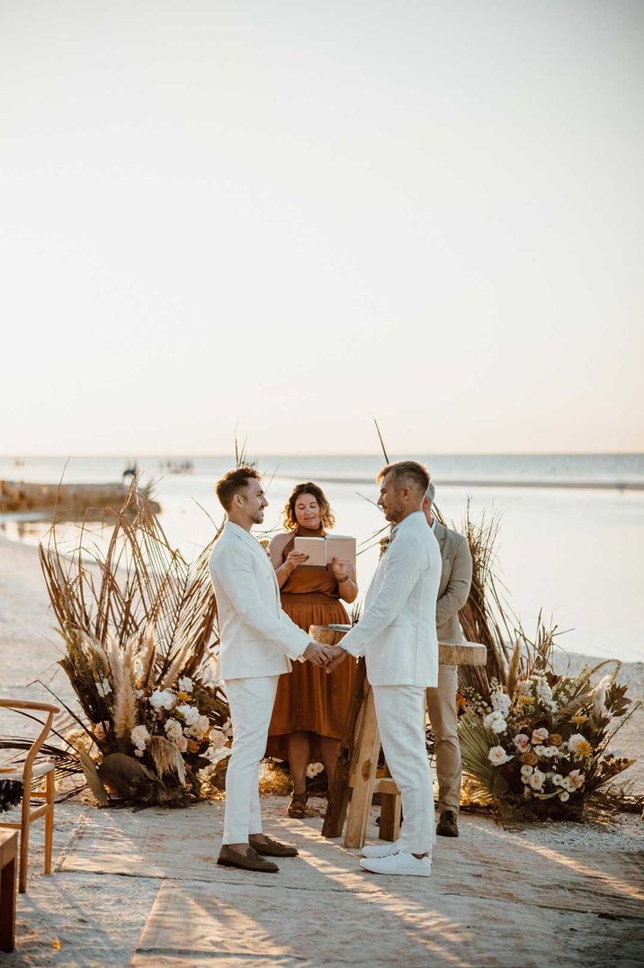 Two grooms in white suits hold hands for their LGBTQI+ wedding ceremony at beach wedding 