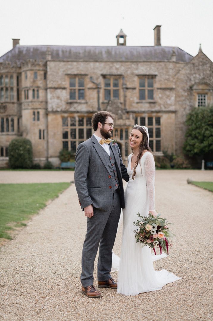 Portrait of the bride in an embellished Eliza Jane Howell wedding dress and the groom in a grey suit standing outside their Brympton House wedding venue 