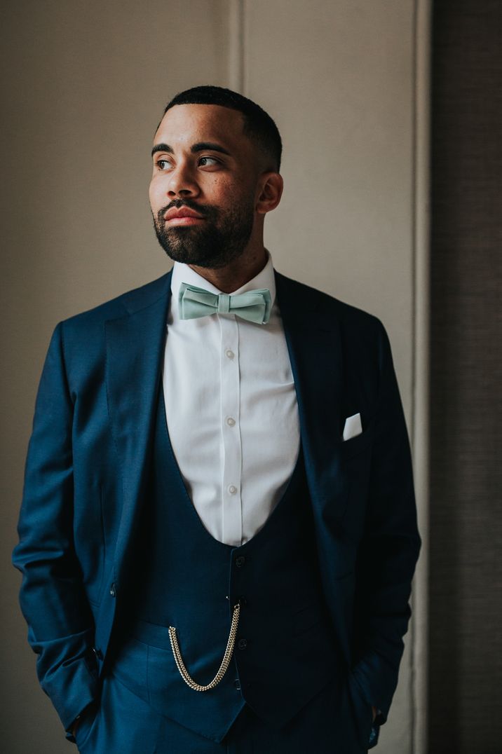 A groom wears a dark blue tuxedo for grooms suit ideas. He wears alight green bow tie. 