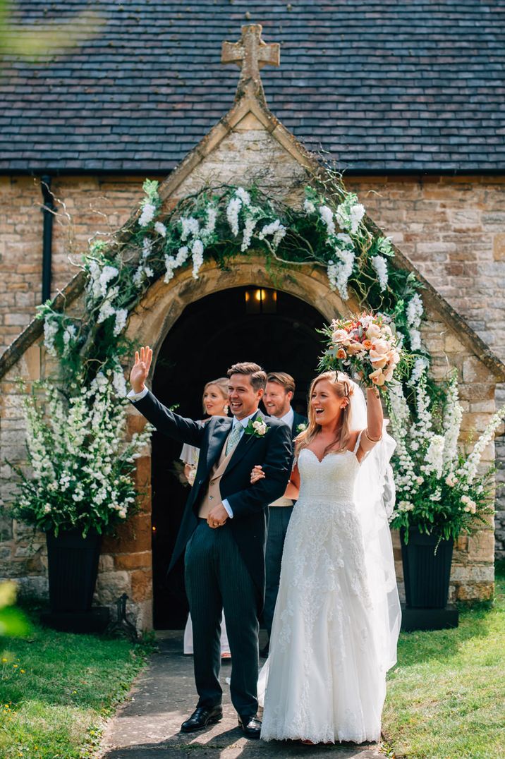 Bride in a strapless lace wedding dress and groom in a morning suit waving outside the church decorated in a white flowers 