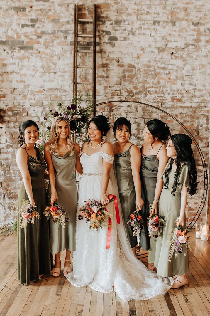 Bridesmaids in sage green satin dresses with bride in off the shoulder lace gown in front of floral moongate 