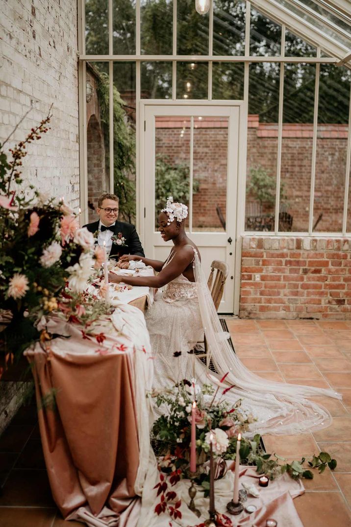 Bride and groom sitting at their luxe wedding tablescape with dust pink tablescape, large florals and tapered candles at Our Beautiful Glasshouse wedding venue 