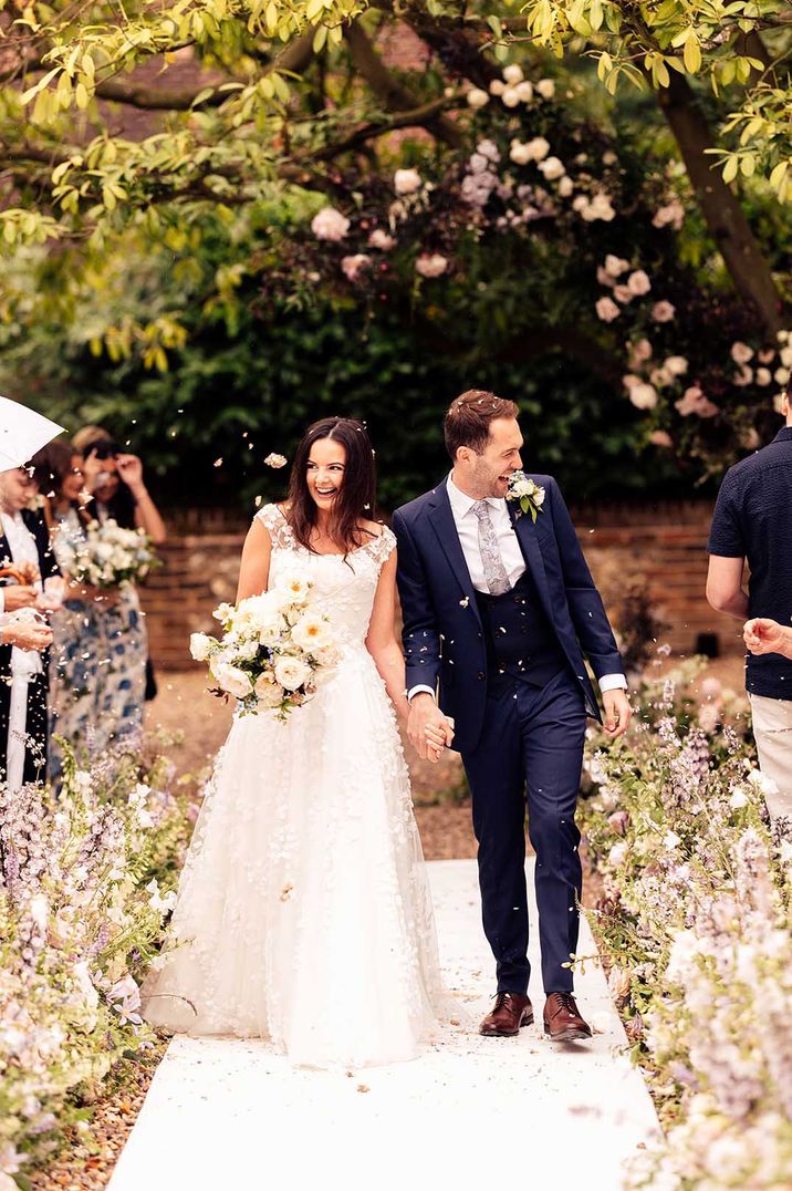 Bride in Phillipa Lepley wedding dress with 3D floral applique with the groom in three piece navy suit with patterned tie walking to confetti exit 