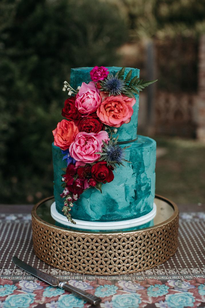 Two tier turquoise wedding cake on a gold cake stand with pink and red peony and rose cake decor