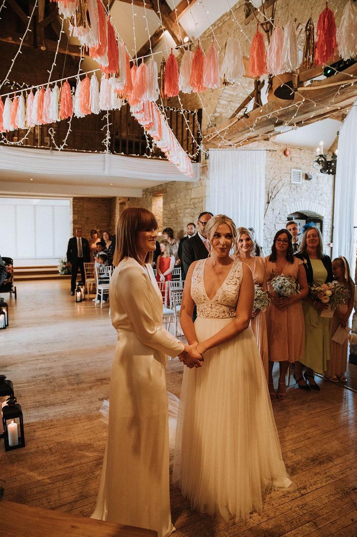 LGBTQI+ wedding ceremony at The Great The Barn with two brides holding hands at the altar as they exchange vows under a canopy of tissue bunting 