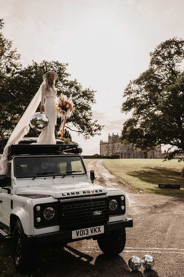 Bride in long veil with disco ball and bouquet on top of a grey Land Rover