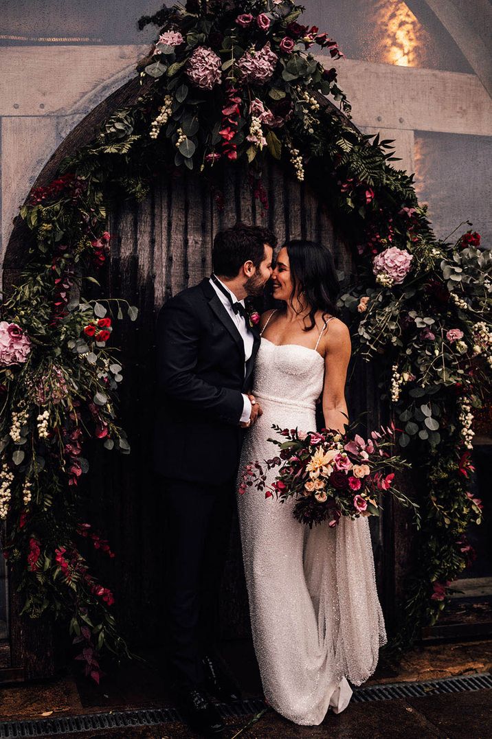 Bride in sparkly beaded wedding dress with the groom underneath pink floral arch decoration 