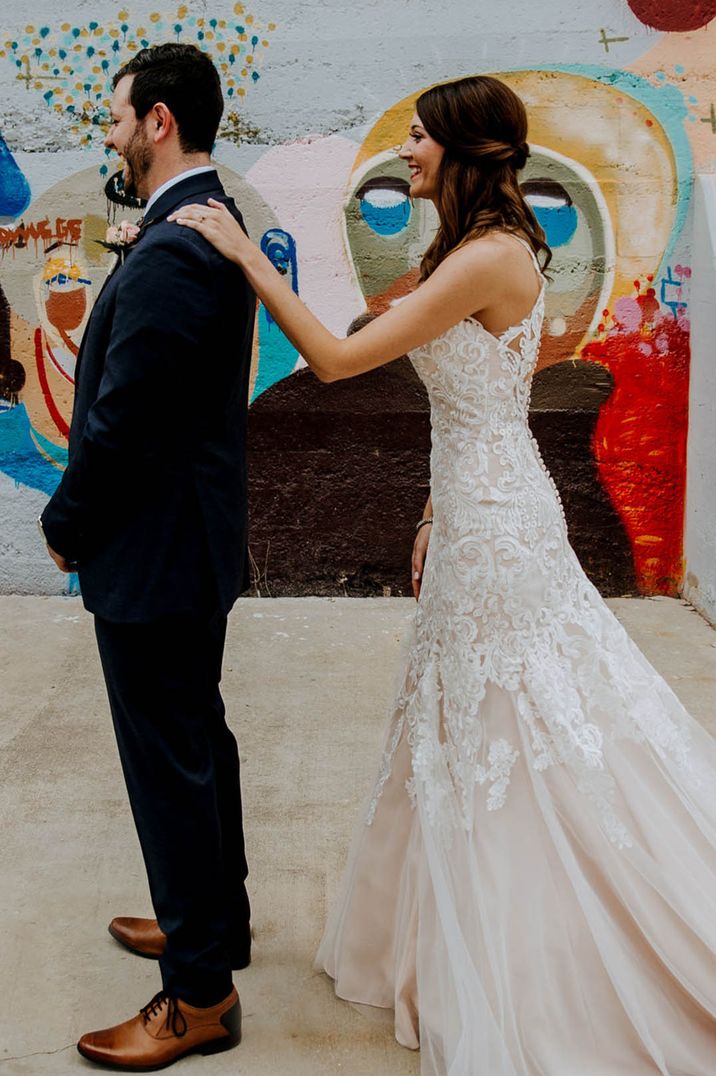 Bride in mermaid wedding dress taps the groom on the shoulder so he can see her in her wedding dress 