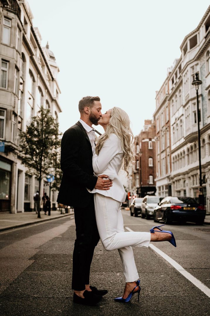 Bride in a trouser suit with blue Manolo Blahnik shoes kicking her heels up as she kisses her groom 