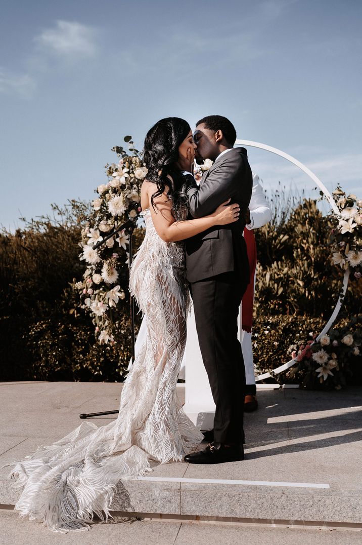 Bride in a couture feather wedding dress kissing her groom in a tuxedo in front on a hoop moon gate altar 