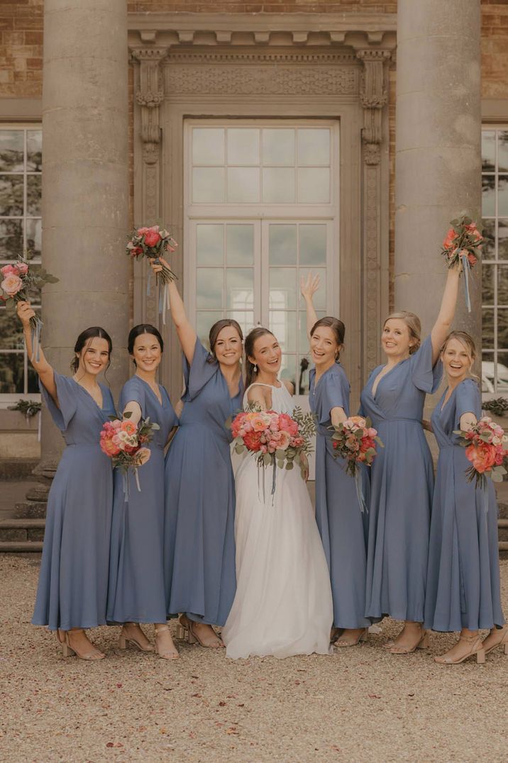 Bride stands with bridesmaids in chiffon blue bridesmaid dresses all holding mixed bouquets at wedding