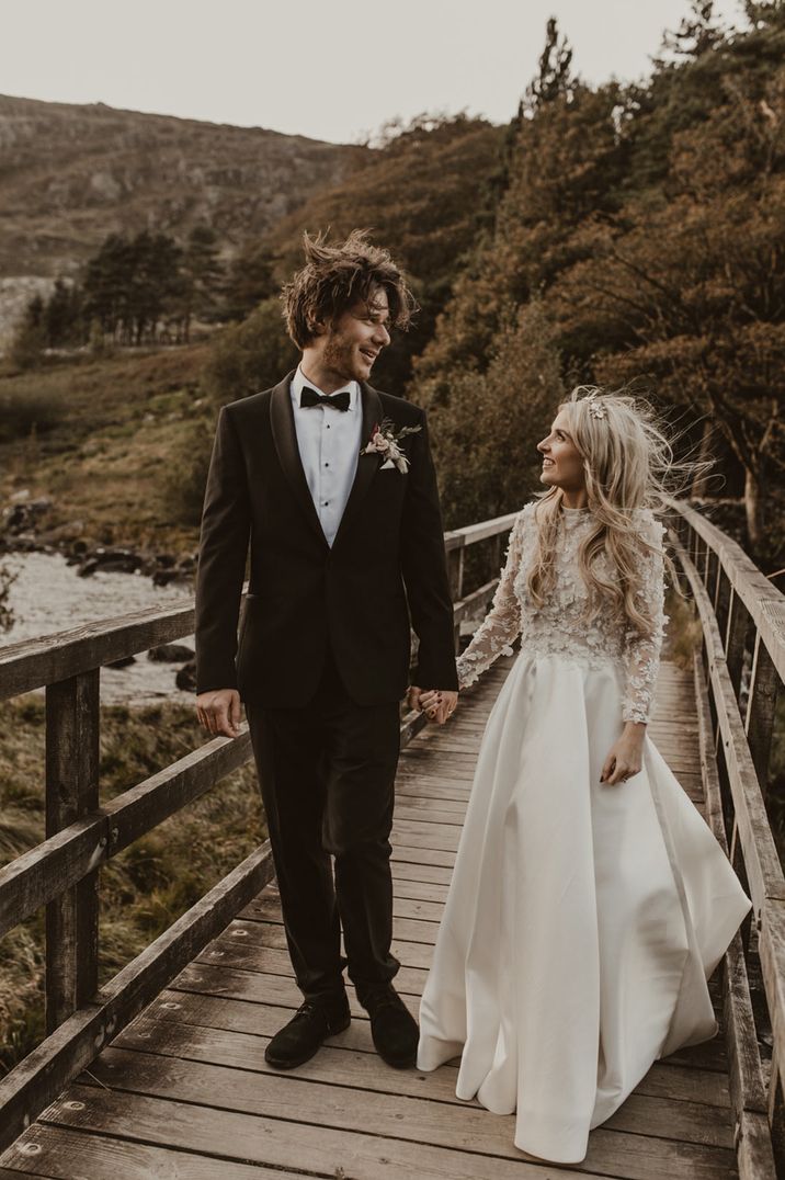 Bride in custom made long sleeve full length wedding dress and groom in dark brown velvet suit standing on a bridge 