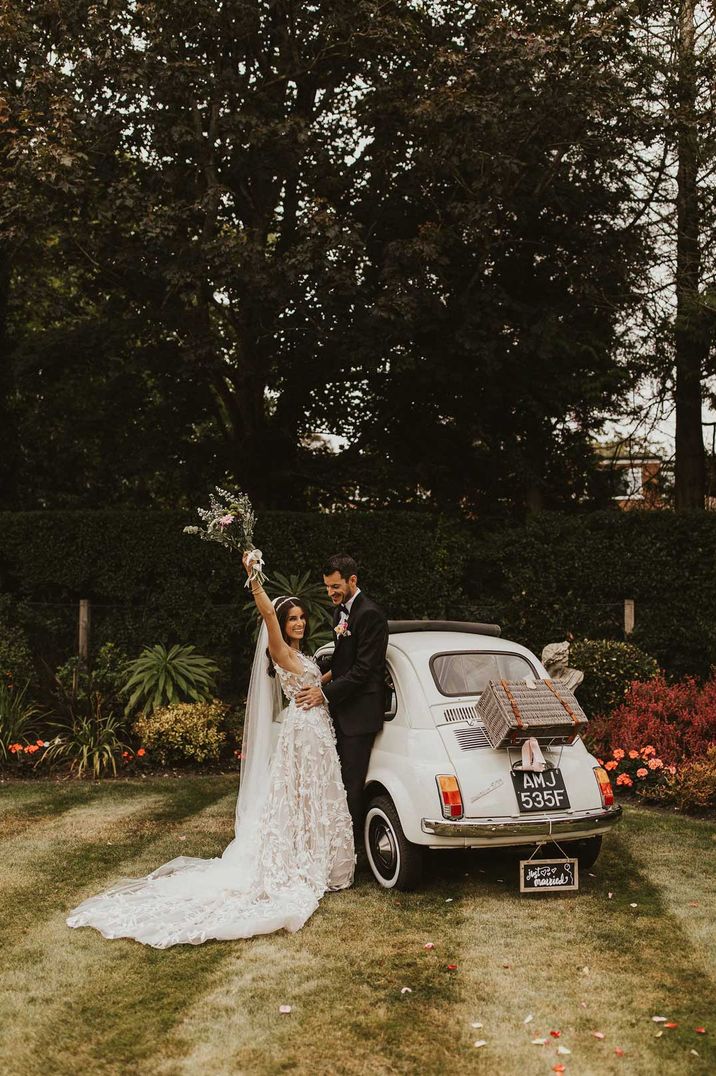 Bride and groom standing beside white Fiat 500 with "Just Married" wedding trunk and suitcase decorations