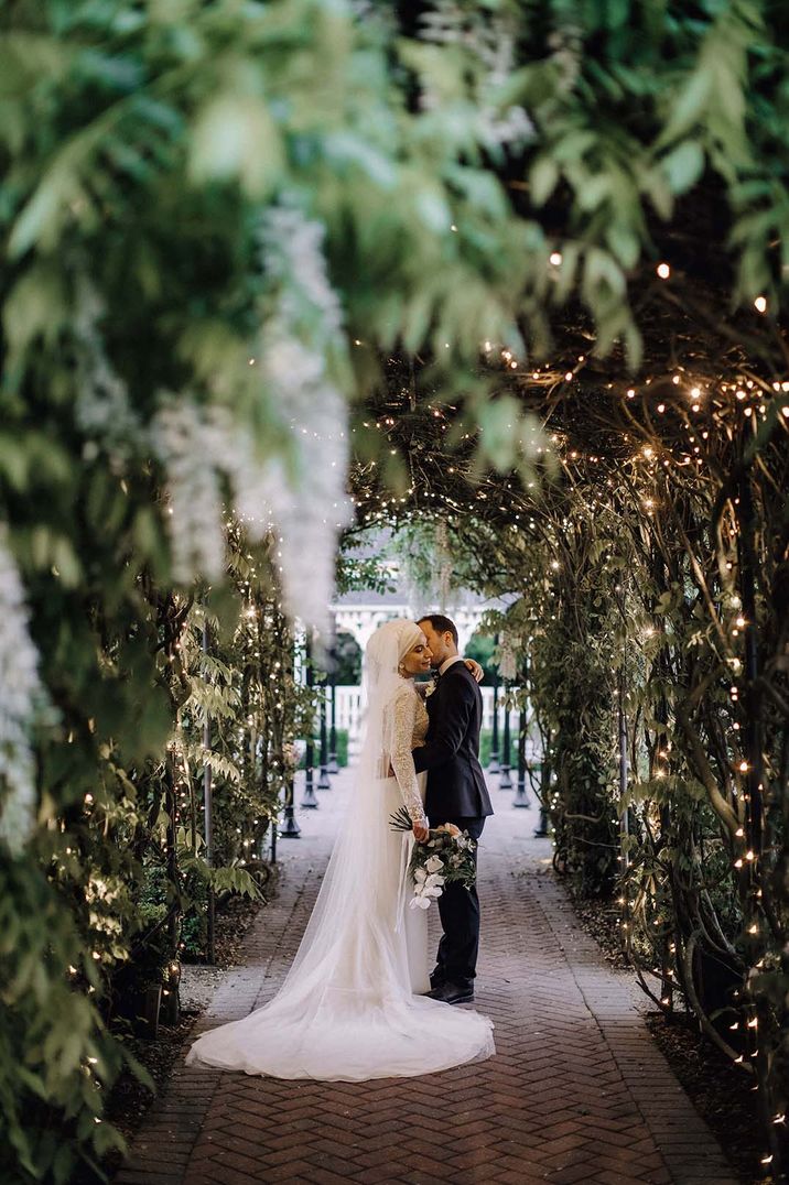 Bride in long sleeve lace wedding dress and groom in dark suit embracing in foliage archway