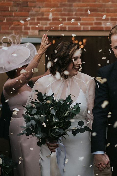 Confetti exit for bride in sheer long sleeve wedding dress walking with the groom holding an all foliage preserved wedding flower bouquet 