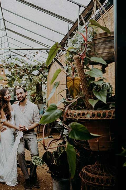 Groom in white shirt and grey trousers and bride in off the shoulder wedding dress holding hands and smiling amidst monstera plant and Chinese money plant, fairy lights and other exotic plants at Petersham Nurseries glasshouse wedding venue 