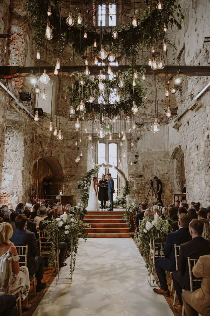 Ceremony room of Lulworth Castle wedding venue - suspended foliage hoops, hanging lightbulbs, wooden beams and large foliage arch at the end of the aisle 