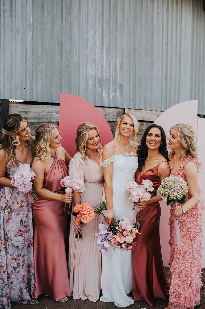 Bride wearing lace off the shoulder wedding dress standing in front of pink wedding screen backdrop with bridesmaids in mismatched bridesmaid dresses