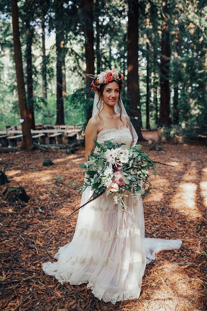 Bride in a sheer wedding dress wearing a flower crown and holding an oversized bouquet at a woodland boho wedding 