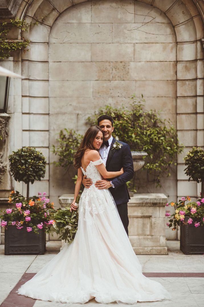 Groom in a navy suit embracing his bride in an off the shoulder Martina Liana wedding dress at Carlton House Terrace wedding venue in London 