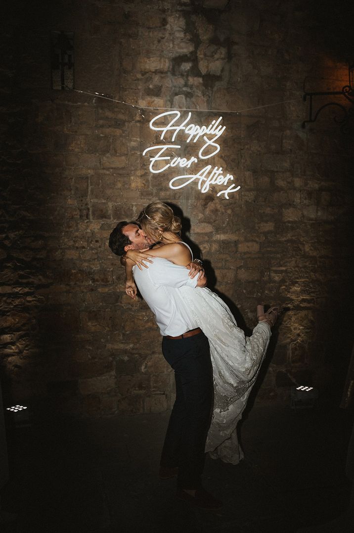 Groom lifts the bride and kisses her in front of a white neon sign 