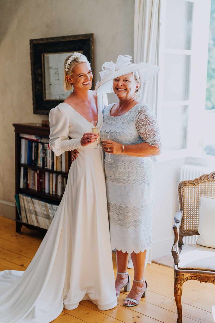 Bride in long sleeve v neck wedding dress standing with mother of the bride in white fascinator and light grey lace dress