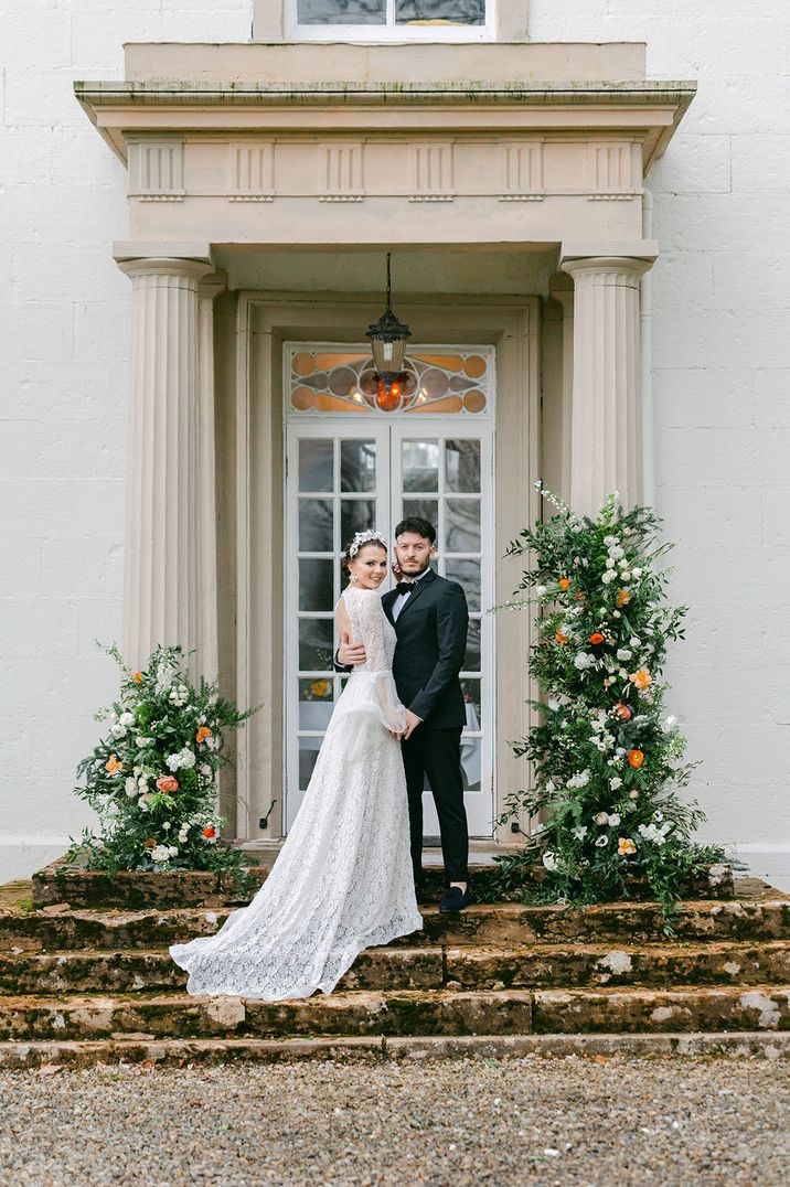 Bride in long sleeve boho lace wedding dress with groom in black tuxedo with orange flower column decorations on the stairs 