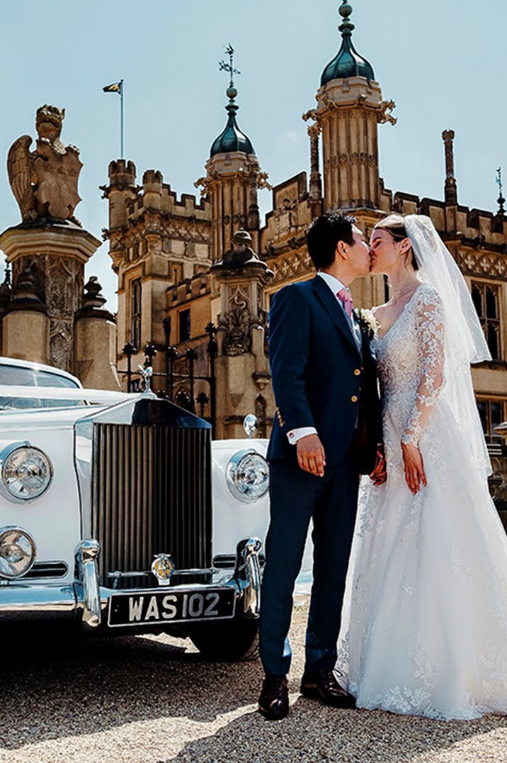 Bride in lace long sleeve wedding dress with groom in black tuxedo with white vintage car at country house wedding venue 