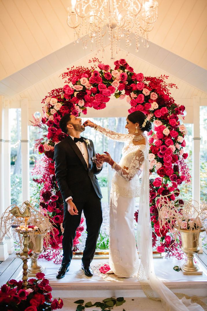 Bride feeding her husband at their multicultural wedding with a bright pink flower arch 