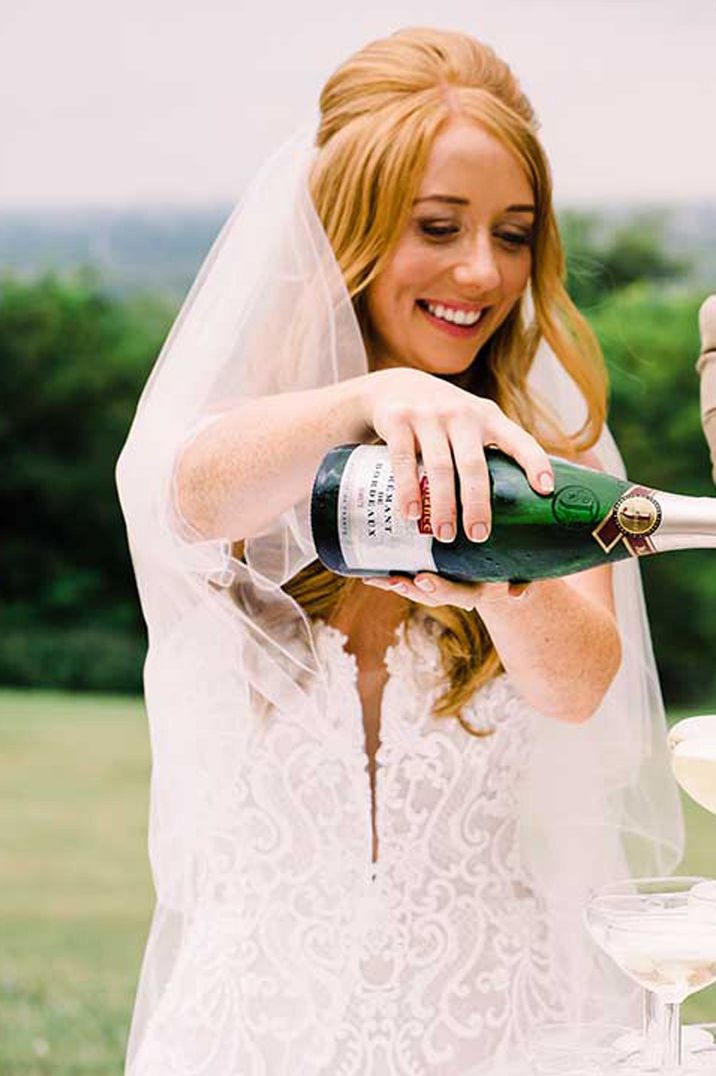 Bride in lace dress and groom in sunglasses and beige suit pouring champagne tower at wedding