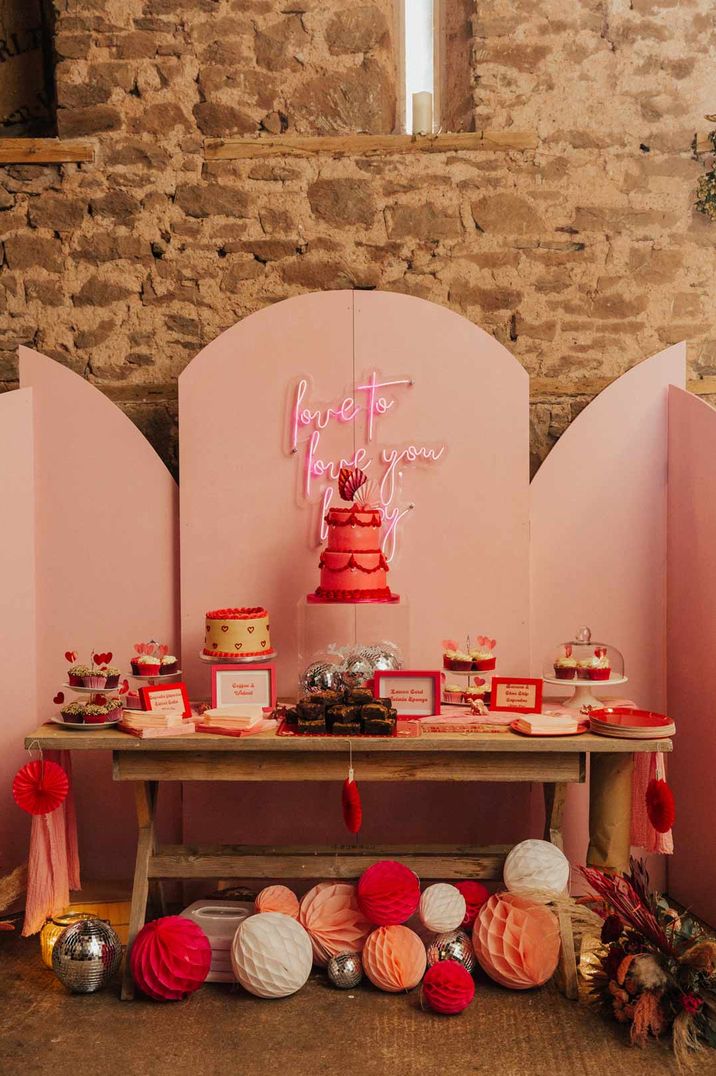 Pink neon sign above the dessert station with two tier hot pink wedding cake, vegan cupcakes and brownies with paper lanterns and disco balls at gender neutral wedding