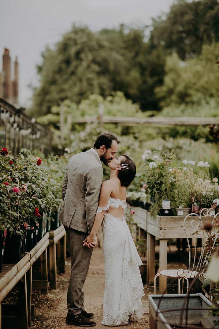 Bride in off the shoulder open back detail wedding dress and groom in grey suit kissing amidst the plants in Petersham Nurseries wedding venue 