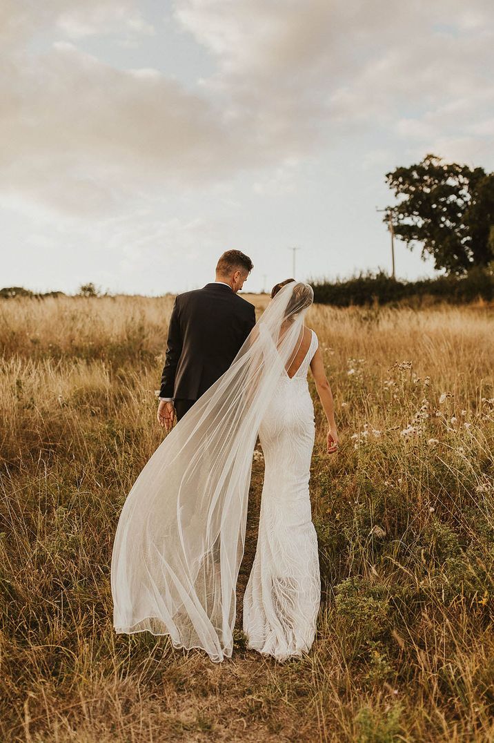 Rustic barn wedding in Bath with the bride in a beaded fitted open back wedding dress and long veil walking hand in hand with the groom 