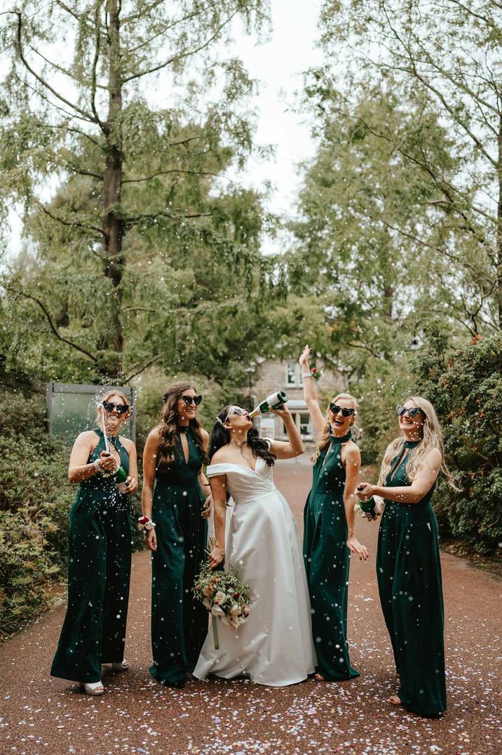 Bride in off shoulder satin wedding dress drinking champagne with bridesmaids in bottle green keyhole bridesmaid dresses with heart shaped sunglasses