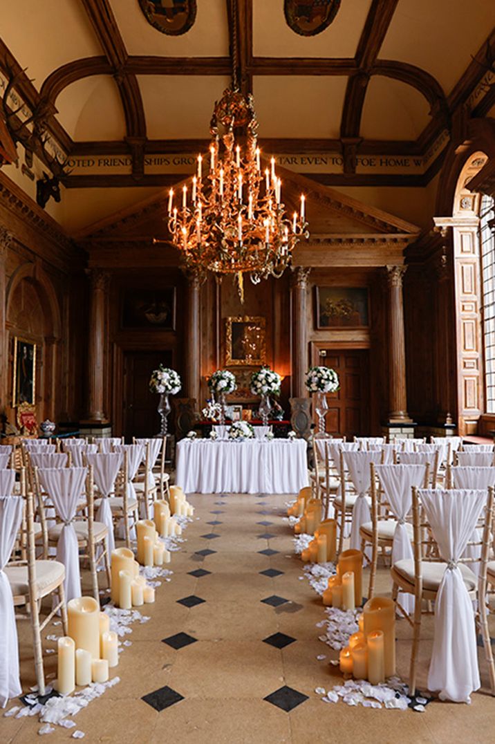 Luxe country house wedding venue in Knebworth House with white fabric chair back decor with chandeliers and candles lining the aisle 